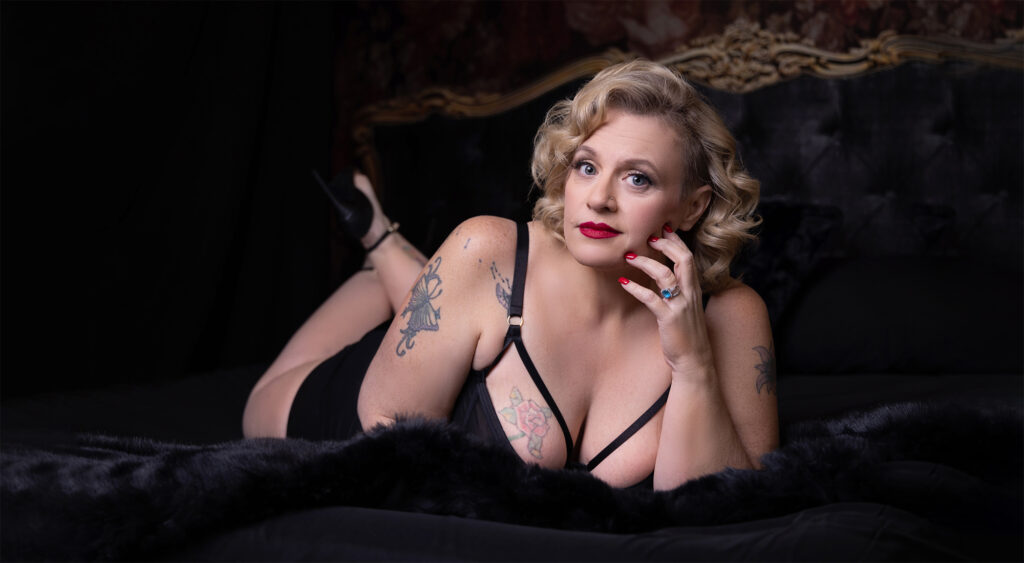 best boudoir photographer near me, this mature woman is laying on a black bed. She has tattoos on her breast and arms.