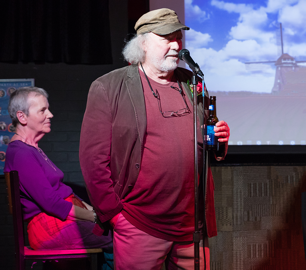 Larry Carius, dressed in maroon shirt with his wife Kay sitting to the side. Larry is holding a beer and speaking into a microphone.