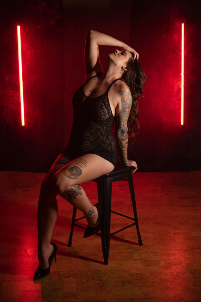 Tattooed woman in black lace bodysuit posing on stool with red studio lights.